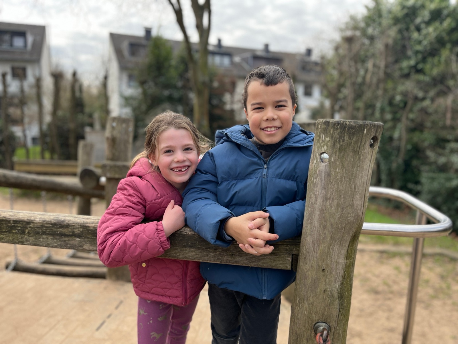 Zwei Kinder halten sich lachend an einem Holzgeländer auf einem Spielplatz fest